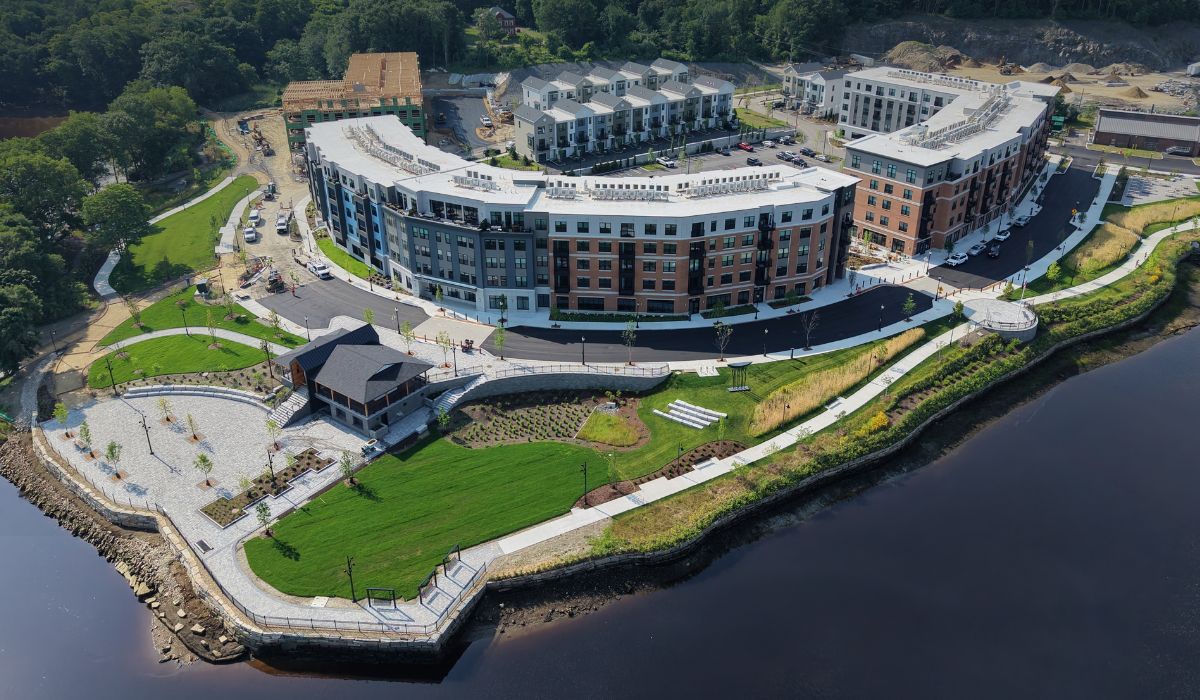 Aerial view of a curved multi‑story residential complex along a waterfront park with green lawns, pathways, and a stone promenade by the water.